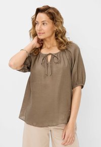 Blusa in lino marrone con scollo arricciato, apertura a chiave frontale e maniche a sbuffo corte. Una leggera lucentezza aggiunge texture. Abbinata a pantaloni chiari.