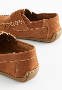 Des chaussures marron en daim à enfiler présentent un bout arrondi avec des détails de couture et une semelle en caoutchouc, avec une texture lisse et un design minimaliste.