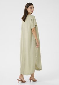 Világoszöld, maxi hosszúságú, rövid ujjú ruha, puha textúrával, oldalsó hasítékokkal és kerek végződéssel. Barna, pántos, magas sarkú szandállal viselve.
