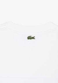 Vue de dos d'une chemise blanche montrant un petit logo de crocodile vert brodé, centré juste sous l'encolure.