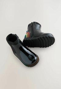 Svarta lackläder ankelskörtar med blixtlås, med en texturerad sula och flerfärgade regnbågsdetaljer på hälen och sidan.