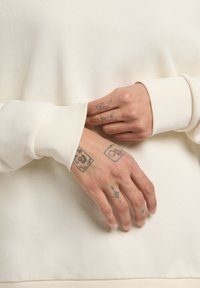 Felpa color crema con una finitura testurizzata, polsini larghi e tatuaggi visibili sulle mani che riposano sul tessuto.