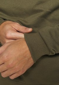 Due mani che aggiustano il polsino di una camicia o maglione a maniche lunghe di colore verde oliva.