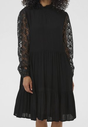 Czarna sukienka do kolan z przezroczystymi koronkowymi długimi rękawami, marszczoną talią i wysokim, falbaniastym kołnierzem, noszona przez osobę o kręconych włosach.