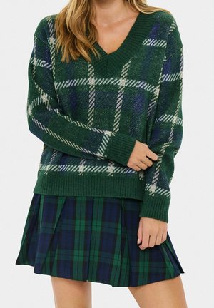 Femme portant un pull à col en V écossais vert et bleu et une jupe plissée écossaise assortie sur fond blanc.
