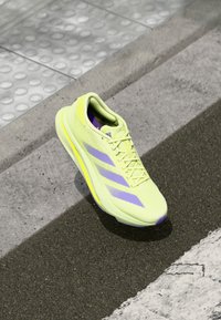 Sneaker verde lime con accenti viola, superiore sintetico leggero, punta rotonda, lacci prominenti e suola testurizzata.
