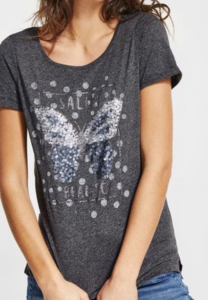 Frau trägt ein dunkelgraues T-Shirt mit einem großen silbernen und blauen Pailletten-Schmetterling und weißen Punkten auf der Vorderseite.