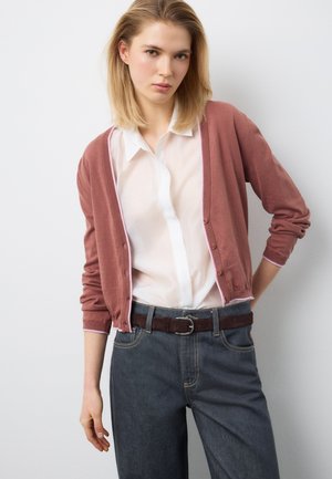 Junge Frau mit glattem blondem Haar, die ein weißes Hemd mit Knöpfen, eine altrosafarbene Strickjacke, dunkle Jeans und einen braunen Gürtel vor weißem Hintergrund trägt.