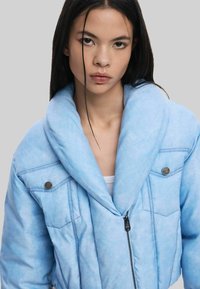 Heledsinine ülekaaluline jakk tekstuuriga viimistlusega. Omab suurt šaalikaelust, lukk sulgemist ja kahte rinna taskut metallaktsentidega.