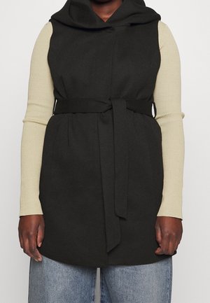 Osoba nosząca czarną bezrękawnikową kamizelkę z paskiem, na beżowym żebrowanym topie z długim rękawem i jasnoniebieskich dżinsach, stojąca na jednolitym tle.