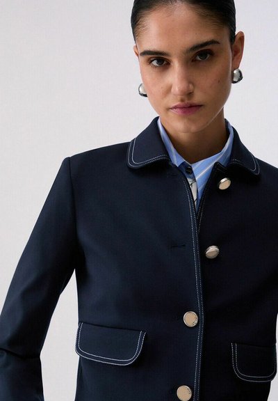 Femme portant un blazer bleu marine avec des surpiqûres blanches contrastantes et des boutons argentés, associé à une chemise bleu clair à col, regardant devant elle.
