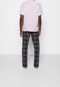 Person wearing a light purple patterned short-sleeve shirt and black pants with purple peace sign prints, standing against a plain background.