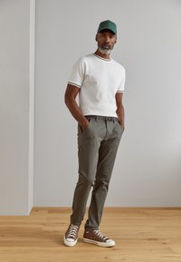 Fehér crew neck póló fekete és szürke csíkokkal, olíva zöld nadrág és barna sportcipő fehér talppal és lyukacsos részletekkel. Zöld sapka.