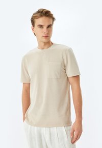 T-shirt beige a maniche corte realizzato in tessuto morbido, con scollatura rotonda e taschino sul petto, indossato con pantaloni in lino di colore chiaro.