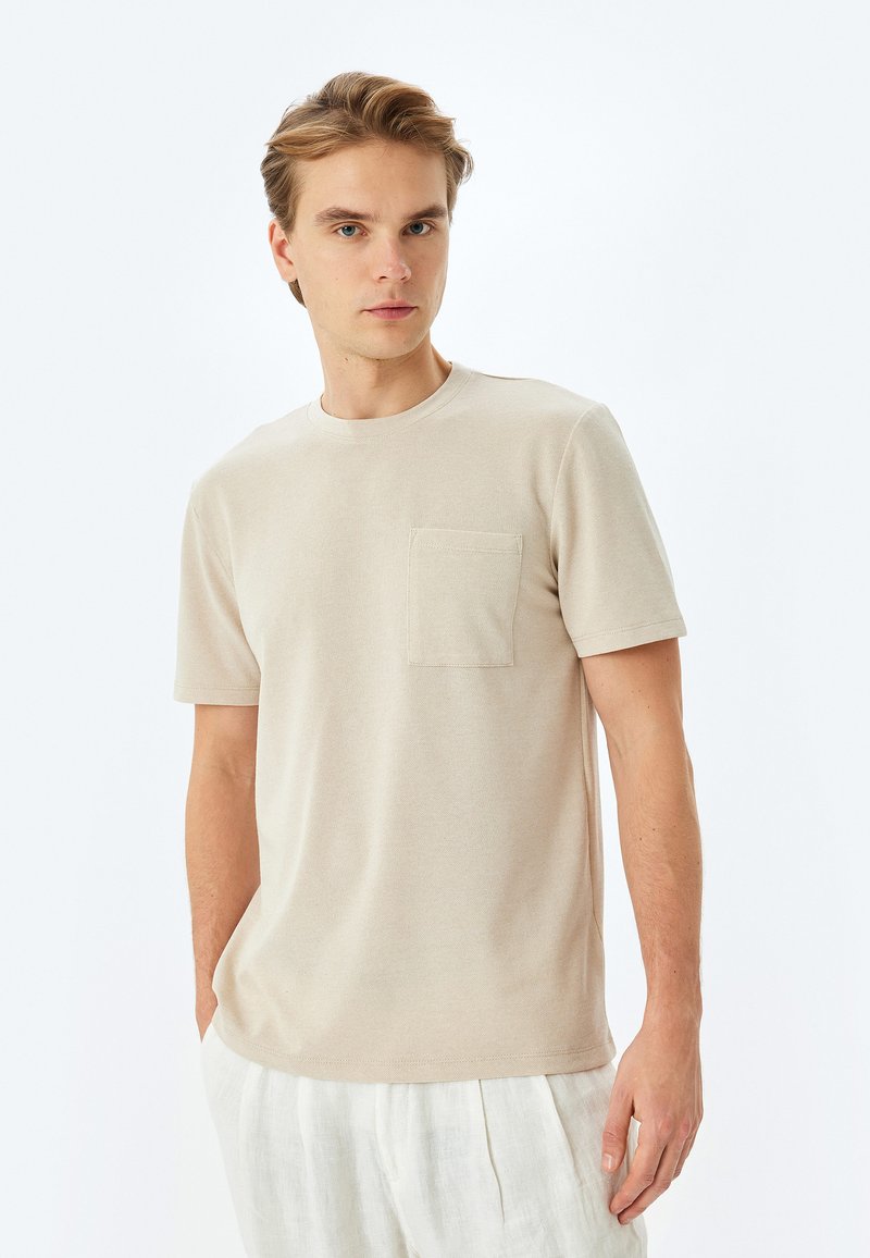 T-shirt beige a maniche corte realizzato in tessuto morbido, con scollatura rotonda e taschino sul petto, indossato con pantaloni in lino di colore chiaro.