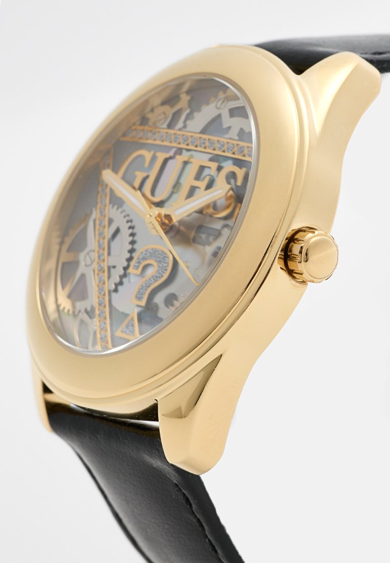 Orologio dorato con quadrante trasparente che mostra gli ingranaggi, cinturino in pelle nera e un prominente logo "GUESS" al centro. Design della cassa rotonda.