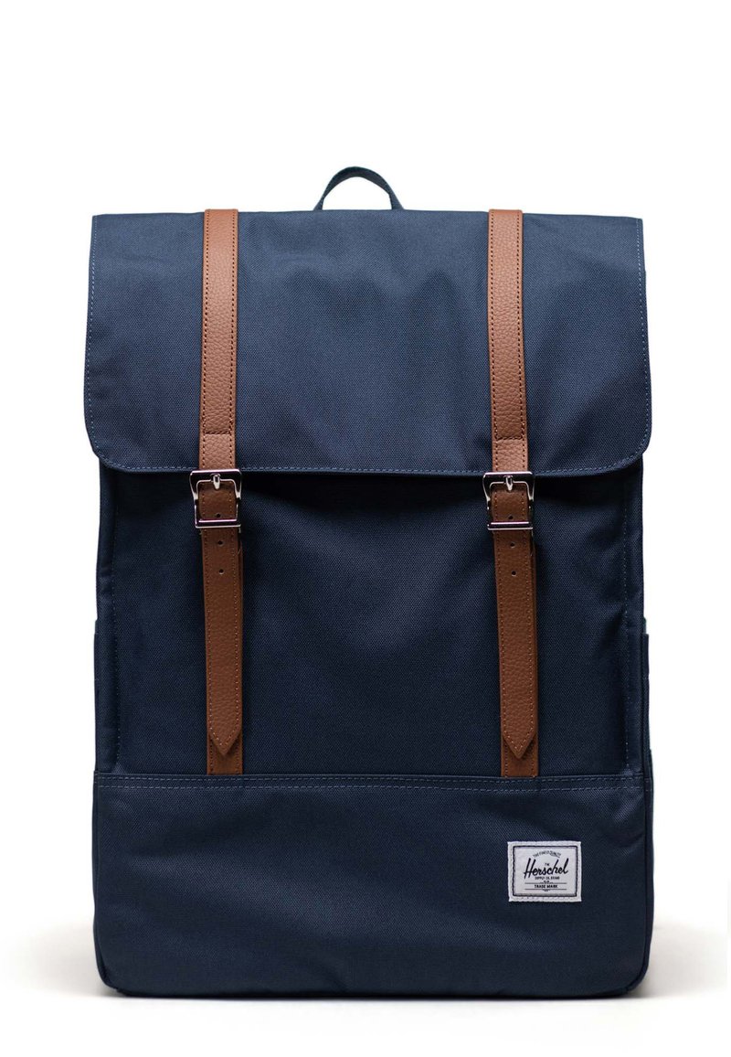 Zaino in tessuto blu navy con spallacci in pelle marrone, chiusura a pattina superiore, forma rettangolare, logo a toppa frontale, superficie texturizzata.