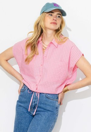 Femme aux longs cheveux blonds portant une chemise rose à manches courtes boutonnée, un jean bleu, une casquette bleue, des bracelets dorés et des colliers superposés.
