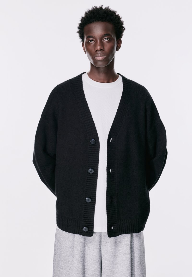 Cardigan tricotat negru cu decolteu în V, cu manșete încrucișate și șase butoane mari. Purtați peste o cămașă albă, asortat cu pantaloni gri.