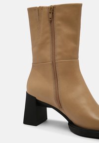 Brązowy skórzany botek za kostkę na słupku, z podwójnym zamkiem błyskawicznym i gładką fakturą. Podeszwa jest czarna, kontrastując z cholewką buta.