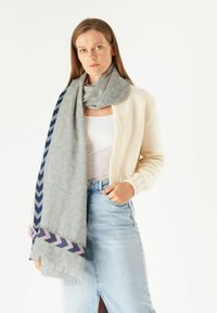 Hellgrauener Schal mit geometrischem Muster, versehen mit marineblauen und rosa Zickzackakzenten, getragen über einer cremefarbenen Strickjacke und einem Jeansrock.