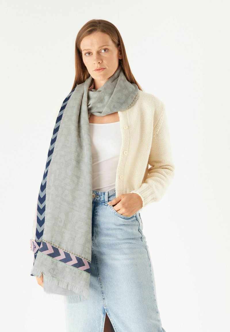 Hellgrauener Schal mit geometrischem Muster, versehen mit marineblauen und rosa Zickzackakzenten, getragen über einer cremefarbenen Strickjacke und einem Jeansrock.