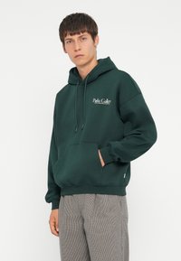 Dunkelgrüner Hoodie mit einer Kängurutasche, Kapuze mit Kordelzug und weißem Textlogo. Trägt man mit hellkarierten Hosen. Weiches Stoffgefühl.