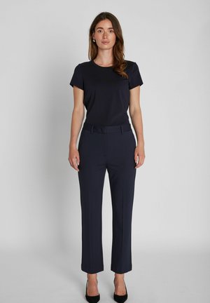Femme debout face à l’avant portant un haut noir à manches courtes, un pantalon bleu marine ajusté avec passants de ceinture, et des talons hauts noirs sur un fond uni.