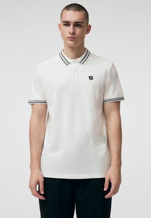 Młody mężczyzna w białej koszulce polo z czarno-białym paskowanym kołnierzykiem i mankietami na rękawach, stojący na jednolitym tle.