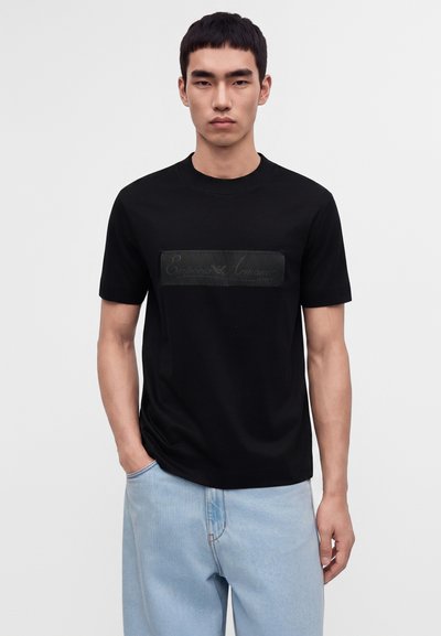 Άνδρας με κοντά μαύρα μαλλιά φοράει μαύρο T-shirt Emporio Armani και ανοιχτό μπλε τζιν, στέκεται με το ένα χέρι στην τσέπη του μπροστά σε απλό φόντο.