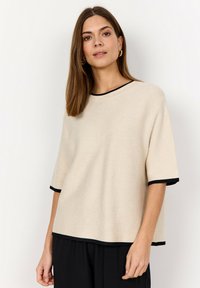 Kvinna med rakt brunt hår, klädd i en beige kortärmad topp med svart kant och svarta byxor, stående mot en enkel vit bakgrund.