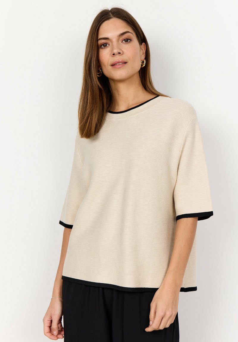 Kvinna med rakt brunt hår, klädd i en beige kortärmad topp med svart kant och svarta byxor, stående mot en enkel vit bakgrund.