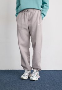 Person iført lyserøde Nike sweatpants og sneakers i pastelfarver, der står på et blåt tæppe med hænderne i lommerne.
