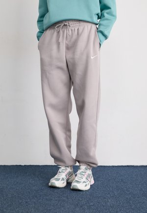 Persona, kas ģērbies gaiši violetos Nike treniņbiksēs un pasteļkrāsās sporta apavos, stāv uz zilas paklāja, rokas ieliktas kabatās.