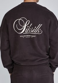 Felpa pullover marrone scuro con una texture morbida. Presenta un grande logo bianco sul retro con testo racchiuso in un design decorativo.
