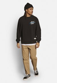 Schwarzer Hoodie mit dem "Good Company"-Logo in Blau und Weiß. Kombiniert mit beigen Hosen und schwarzen Sneakern, die weiße Sohlen und Schnürsenkel haben.