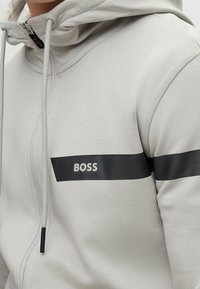 Ljust grå hoodie i mjukt material, med en svart horisontell rand som har "BOSS"-logotypen, framzipper och justerbar dragsko på huvan.
