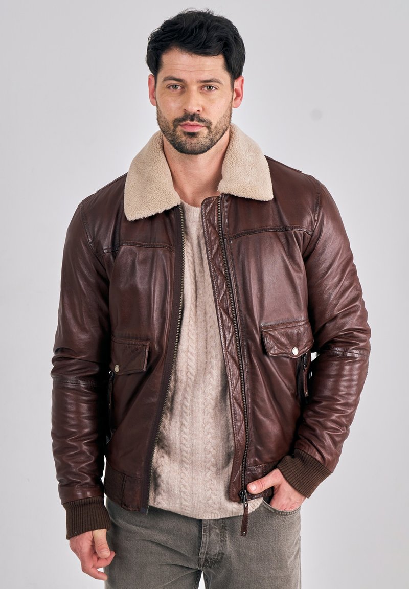 Man wearing a brown leather jacket with a beige shearling collar over a light knit sweater and gray pants, standing against a plain background.