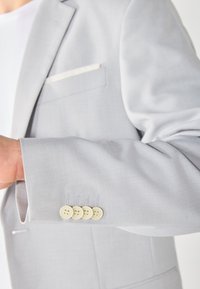 Hellgrauer Blazer mit glatter Textur, ausgestattet mit einer Tasche mit weißem Saum und vier cremefarbenen Knöpfen am Bund. Maßgeschneiderter Schnitt, moderne Passform.