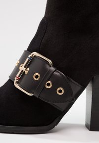 Bottine en daim noir à talon épais, avec une large sangle en cuir noir munie d'une boucle dorée et d'œillets métalliques.