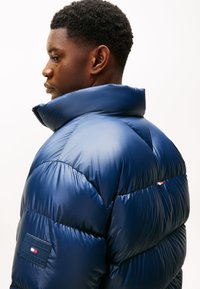Ung mand i en højhalset, skinnende navy blå puffjakke med Tommy Hilfiger-logo på venstre ærme, vendt mod venstre foran en ensfarvet baggrund.