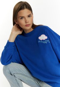 Felpa blu con scollo rotondo, maniche lunghe e dettaglio ricamato di una nuvola. Presenta il testo "nuvoloso fuori, felice dentro" in colori a contrasto.