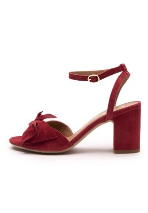 Czerwony zamszowy sandał na wysokim słupku z szerokim obcasem, otwartym noskiem i kokardką z przodu. Regulowany pasek wokół kostki ze złotym okuciem.