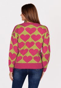 Vrouw met lang bruin haar, gekleed in een roze en groene trui met grote roze harten, in combinatie met blauwe jeans, van achteren weergegeven.