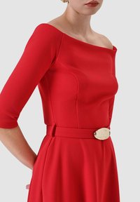 Abito aderente rosso con scollatura a spalle scoperte, maniche a tre quarti e vita con cintura caratterizzata da un dettaglio decorativo con fibbia metallica.