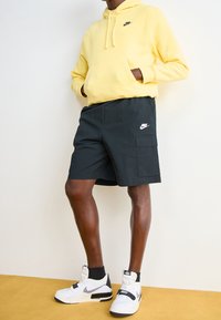 Felpa gialla Nike, pantaloni cargo neri e sneakers bianche con dettagli neri. Design con cappuccio, tasche sui pantaloni e dettagli del logo visibili.