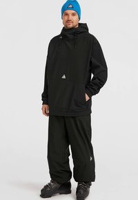 Giacca da sci nera con cappuccio, dotata di tasca frontale e coulisse regolabili. Abbinata a pantaloni neri larghi e un berretto azzurro lavorato a maglia.