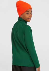 Groen overhemd met lange mouwen en een hoge kraag, gecombineerd met een oranje geribde beanie. De stof lijkt soepel en geschikt voor actieve kleding.