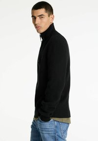 Schwarzer gerippter Strickpullover mit hohem Kragen und halbem Reißverschluss, über einem olivgrünen Hemd getragen, kombiniert mit hellblauen Jeans.