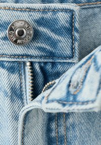 Gros plan sur un tissu en denim bleu clair avec un bouton en argent gravé de "AUTHENTIC DENIM" et une fermeture éclair métallique partiellement visible.